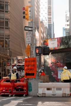 A busy urban street scene with construction work in progress. Traffic lights are overhead, and various street signs are positioned, including a 'sidewalk closed, please use other side' sign. Two construction workers, one in a yellow vest and another with a security jacket, are visible. The area is cordoned off with red and white barriers, and there is a building in the background with a colorful mural.