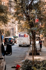 A city street scene with a FedEx delivery truck in the center, surrounded by other vehicles. Trees with green leaves line the sidewalk, and a person walks along the sidewalk. A red no standing parking sign is visible above the street, and buildings with large windows can be seen in the background.