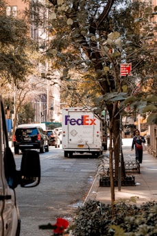 A city street scene with a FedEx delivery truck in the center, surrounded by other vehicles. Trees with green leaves line the sidewalk, and a person walks along the sidewalk. A red no standing parking sign is visible above the street, and buildings with large windows can be seen in the background.