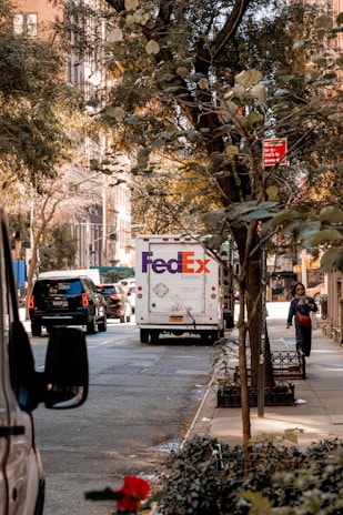 A city street scene with a FedEx delivery truck in the center, surrounded by other vehicles. Trees with green leaves line the sidewalk, and a person walks along the sidewalk. A red no standing parking sign is visible above the street, and buildings with large windows can be seen in the background.