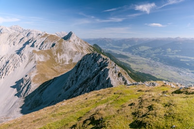 A breathtaking alpine landscape featuring a range of rugged mountains with steep slopes and prominent ridges. The foreground shows a grassy, rocky terrain leading to the edge of the mountains. In the distance, a wide valley with villages and lush greenery is visible under a clear blue sky.
