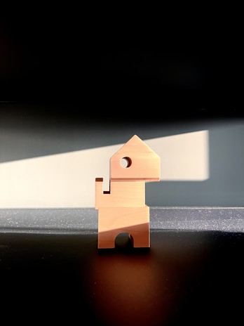 A wooden block shaped like a simplified, abstract figure stands upright on a dark surface. The design resembles a combination of geometric shapes with a square body, circular head, and triangular top. The light casts dramatic shadows around the object, highlighting its distinct contours.