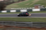 A black car is racing on a track with flames visibly emanating from the rear exhaust, indicating high speed. The surroundings include blurred green grass and white and green barriers, suggesting motion and a racing environment.