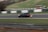 A black car is racing on a track with flames visibly emanating from the rear exhaust, indicating high speed. The surroundings include blurred green grass and white and green barriers, suggesting motion and a racing environment.