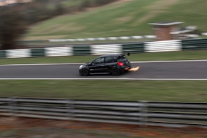 A black car is racing on a track with flames visibly emanating from the rear exhaust, indicating high speed. The surroundings include blurred green grass and white and green barriers, suggesting motion and a racing environment.