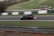 A black car is racing on a track with flames visibly emanating from the rear exhaust, indicating high speed. The surroundings include blurred green grass and white and green barriers, suggesting motion and a racing environment.