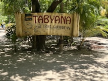 A wooden sign with the text 'TABYANA' and 'THE LOST CARIBBEAN' is displayed. The sign is set against a backdrop of lush green foliage, including palm trees. The ground is sandy, suggesting a tropical or beach setting.
