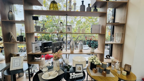 A display of various decorative items arranged on wooden shelves and tables, set against a large window. The items include vases of different sizes and colors, framed pictures, artificial flowers, ceramic teapots, and books. The background features a view of a street with trees in bloom and cars passing by.