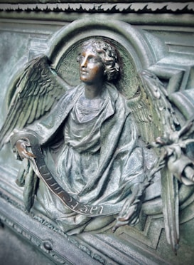 A detailed sculpture of an angel is depicted, featuring intricate wings and a serene expression. The angel is holding a scroll with inscriptions and appears to be carved from metal with a patina finish, giving it an antique and sacred appearance.