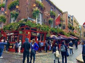 A lively street scene featuring a corner pub named 'The Temple Bar,' adorned with hanging baskets of lush green plants and flowers. People are milling about, some casually dressed and others taking photos. The pub exterior is a vibrant red with multiple signs and banners, and the cobblestone street adds a classic touch. Lamp posts are visible, adding to the quaint atmosphere.