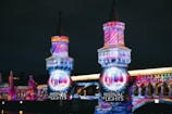 A night scene featuring a bridge illuminated with vibrant, colorful light projections in artistic patterns. The words 'Festival of Lights' are prominently displayed, highlighting the event. The bridge's architectural details are accentuated by the intricate light designs.