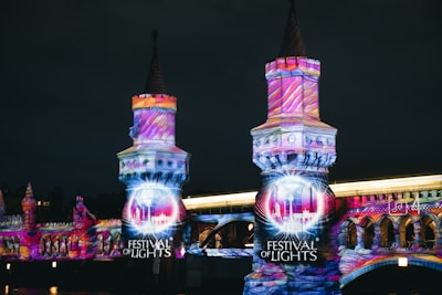 A night scene featuring a bridge illuminated with vibrant, colorful light projections in artistic patterns. The words 'Festival of Lights' are prominently displayed, highlighting the event. The bridge's architectural details are accentuated by the intricate light designs.