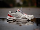 A white and red athletic shoe is positioned on a reflective surface, showing a clear reflection of the shoe. The shoe has a unique textured design with visible branding and design elements on its side. The background is slightly blurred, giving emphasis to the shoe and its reflection.