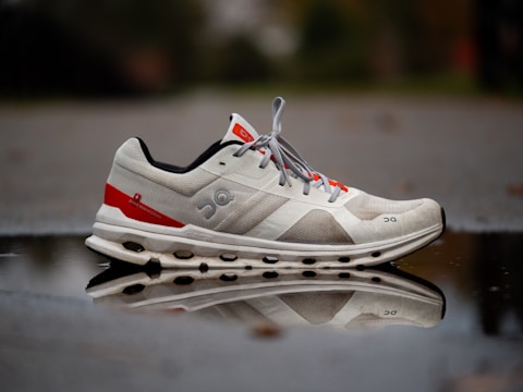 A white and red athletic shoe is positioned on a reflective surface, showing a clear reflection of the shoe. The shoe has a unique textured design with visible branding and design elements on its side. The background is slightly blurred, giving emphasis to the shoe and its reflection.