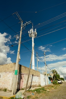 A street scene featuring utility poles with numerous power lines against a clear blue sky. A tall telecommunications tower is visible, surrounded by a few buildings with graffiti on their walls. The street is paved with cobblestones and bordered by grassy patches and scattered litter.