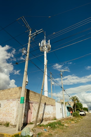 A street scene featuring utility poles with numerous power lines against a clear blue sky. A tall telecommunications tower is visible, surrounded by a few buildings with graffiti on their walls. The street is paved with cobblestones and bordered by grassy patches and scattered litter.
