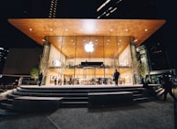 An Apple Store with a modern design featuring a large glass facade and a wooden overhanging roof. The store is illuminated against the night sky, with people visible both inside and outside. Trees with subtle lighting flank the entrance.