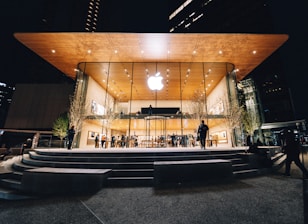 An Apple Store with a modern design featuring a large glass facade and a wooden overhanging roof. The store is illuminated against the night sky, with people visible both inside and outside. Trees with subtle lighting flank the entrance.