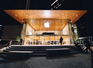 An Apple Store with a modern design featuring a large glass facade and a wooden overhanging roof. The store is illuminated against the night sky, with people visible both inside and outside. Trees with subtle lighting flank the entrance.