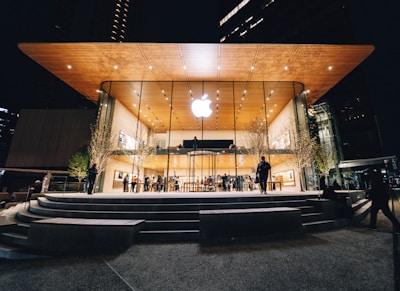 An Apple Store with a modern design featuring a large glass facade and a wooden overhanging roof. The store is illuminated against the night sky, with people visible both inside and outside. Trees with subtle lighting flank the entrance.