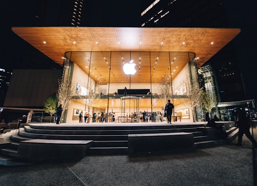 An Apple Store with a modern design featuring a large glass facade and a wooden overhanging roof. The store is illuminated against the night sky, with people visible both inside and outside. Trees with subtle lighting flank the entrance.