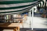 A close-up view of a green and white striped awning with a string of light bulbs attached underneath. In the blurred background, wooden tables and benches are visible, suggesting an outdoor seating area possibly at a cafe or restaurant.