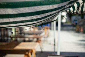 A close-up view of a green and white striped awning with a string of light bulbs attached underneath. In the blurred background, wooden tables and benches are visible, suggesting an outdoor seating area possibly at a cafe or restaurant.