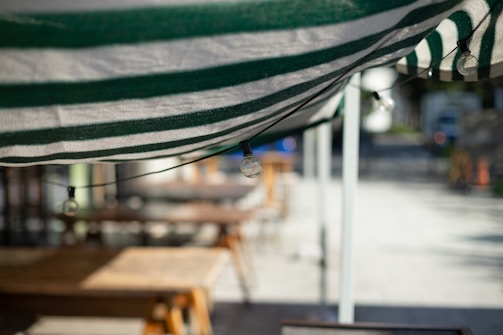 A close-up view of a green and white striped awning with a string of light bulbs attached underneath. In the blurred background, wooden tables and benches are visible, suggesting an outdoor seating area possibly at a cafe or restaurant.