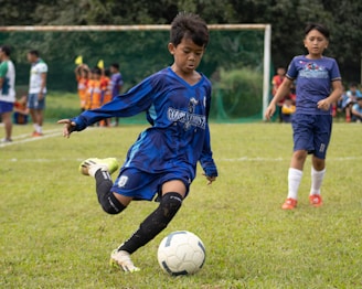 A young boy in a blue soccer jersey is actively engaged in kicking a soccer ball on a grassy field. His focused expression and body posture suggest determination and skill. Other children, some dressed in soccer uniforms, are visible in the background, indicating a soccer game or practice. The scene is set with green grass, a goalpost, and several spectators or teammates on the sidelines.