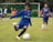 A young boy in a blue soccer jersey is actively engaged in kicking a soccer ball on a grassy field. His focused expression and body posture suggest determination and skill. Other children, some dressed in soccer uniforms, are visible in the background, indicating a soccer game or practice. The scene is set with green grass, a goalpost, and several spectators or teammates on the sidelines.