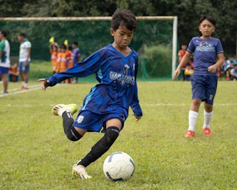 A young boy in a blue soccer jersey is actively engaged in kicking a soccer ball on a grassy field. His focused expression and body posture suggest determination and skill. Other children, some dressed in soccer uniforms, are visible in the background, indicating a soccer game or practice. The scene is set with green grass, a goalpost, and several spectators or teammates on the sidelines.