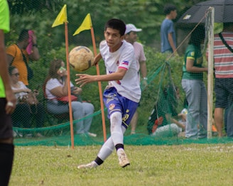 A soccer player in a white and blue uniform is seen in action on the field, focusing intently on controlling the ball with precision. Behind him, spectators watch from the sidelines, seated or standing near a net barrier. The setting indicates an outdoor event on a grassy pitch with vibrant green surroundings.