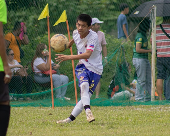 A soccer player in a white and blue uniform is seen in action on the field, focusing intently on controlling the ball with precision. Behind him, spectators watch from the sidelines, seated or standing near a net barrier. The setting indicates an outdoor event on a grassy pitch with vibrant green surroundings.