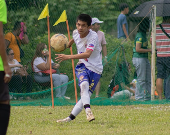 A soccer player in a white and blue uniform is seen in action on the field, focusing intently on controlling the ball with precision. Behind him, spectators watch from the sidelines, seated or standing near a net barrier. The setting indicates an outdoor event on a grassy pitch with vibrant green surroundings.