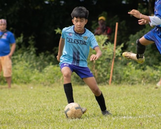 A young soccer player in a light blue jersey is engaged in a game on a grassy field. The player is focused on controlling the soccer ball with his legs. There are spectators in the background, watching the game.