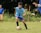 A young soccer player in a light blue jersey is engaged in a game on a grassy field. The player is focused on controlling the soccer ball with his legs. There are spectators in the background, watching the game.