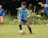 A young soccer player in a light blue jersey is engaged in a game on a grassy field. The player is focused on controlling the soccer ball with his legs. There are spectators in the background, watching the game.
