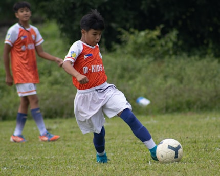 Two young soccer players in orange jerseys and white shorts are on a grassy field. One player is intensely focused as they kick a white and black soccer ball, while the other player watches from behind. The background consists of blurred greenery, suggesting an outdoor setting.