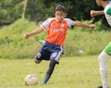 A young soccer player wearing an orange jersey with navy shorts is actively kicking a soccer ball on a grassy field. The player appears focused and is captured mid-action with one leg extended towards the ball. There is another player partially visible in the background.
