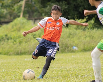 A young soccer player wearing an orange jersey with navy shorts is actively kicking a soccer ball on a grassy field. The player appears focused and is captured mid-action with one leg extended towards the ball. There is another player partially visible in the background.