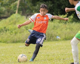 A young soccer player wearing an orange jersey with navy shorts is actively kicking a soccer ball on a grassy field. The player appears focused and is captured mid-action with one leg extended towards the ball. There is another player partially visible in the background.