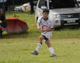 A young soccer player in a white and blue uniform is actively playing on a grassy field, focusing on the ball mid-air. A white vehicle and a red umbrella are visible in the background.