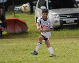 A young soccer player in a white and blue uniform is actively playing on a grassy field, focusing on the ball mid-air. A white vehicle and a red umbrella are visible in the background.