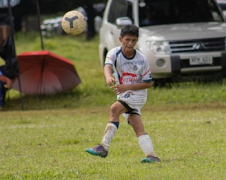 A young soccer player in a white and blue uniform is actively playing on a grassy field, focusing on the ball mid-air. A white vehicle and a red umbrella are visible in the background.
