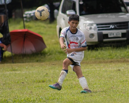 A young soccer player in a white and blue uniform is actively playing on a grassy field, focusing on the ball mid-air. A white vehicle and a red umbrella are visible in the background.