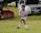 A young soccer player in a white and blue uniform is actively playing on a grassy field, focusing on the ball mid-air. A white vehicle and a red umbrella are visible in the background.