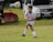 A young soccer player in a white and blue uniform is actively playing on a grassy field, focusing on the ball mid-air. A white vehicle and a red umbrella are visible in the background.