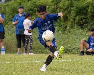 A young soccer player in a blue uniform kicks a ball on a grassy field, with spectators in the background. The action suggests an intense focus and dynamic movement, while other players and onlookers add depth to the scene.