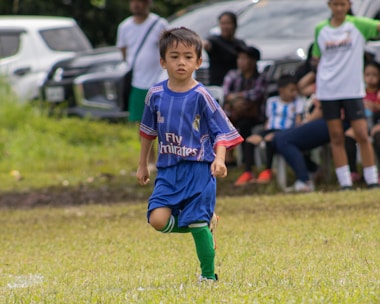 A young child wearing a blue soccer jersey is playing on a grassy field. People are gathered in the background, some seated and others standing, with parked cars visible behind them.