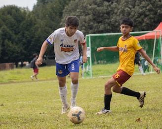 Two young soccer players in action on a grassy field, focused on controlling the ball. One wears a white and blue uniform while the other wears a yellow and red uniform. In the background, a soccer goalpost and trees are visible.